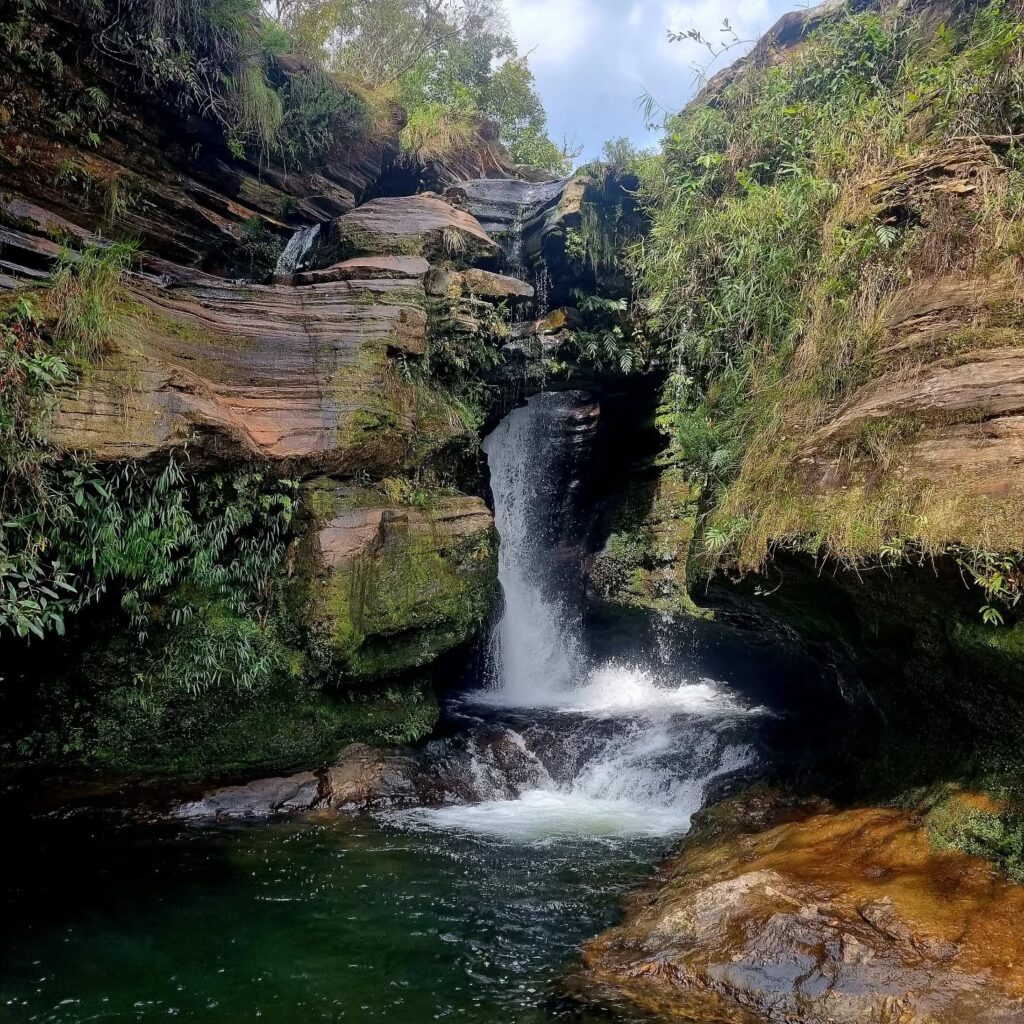 Vista frontal da Cachoeira da Pedra Furada, com água caindo por uma abertura na rocha, formando um poço cercado por vegetação nativa.