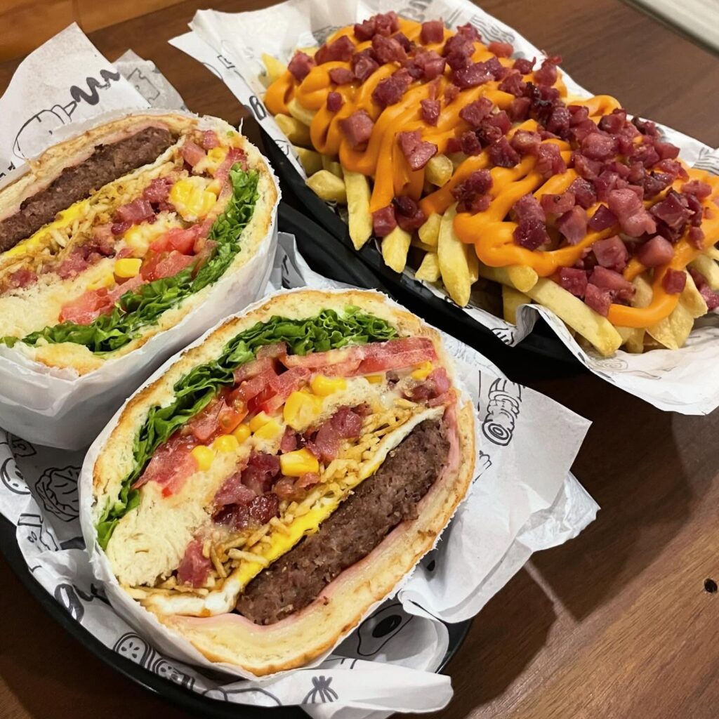 Hambúrguer artesanal suculento acompanhado de uma porção de batatas fritas com queijo derretido e bacon crocante – combinação irresistível do Chapa Quente Luminárias.