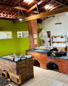Interior do restaurante com fogão a lenha, comida servida em panelas e ambiente rústico e acolhedor.