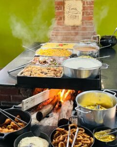 Comida caseira servida no fogão a lenha, com pratos variados e fumaça saindo das panelas.